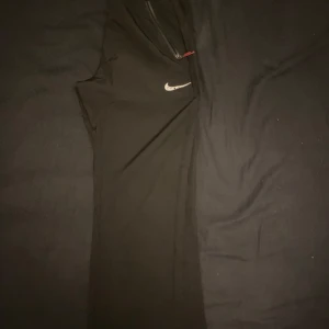 Svarta träningsbyxor från Nike - Snygga svarta träningsbyxor från Nike med vita detaljer och snörning i midjan. Funkar bra vid träning och är använda ett par gånger men är i ett mycket bra skick