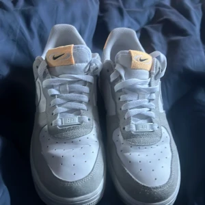 Nike Air Force i vitt och grått - Snygga Nike Air Force sneakers i vitt och grått med klassisk design. Skorna har en vit bas med gråa detaljer och en liten orange Nike-logga på plösen. Perfekta för en stilren look. Storlek 39 Rengör inna beställning