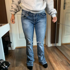 Blå jeans med broderade detaljer - Snygga blå jeans med slitningar och broderade detaljer på bakfickorna. Jeansen har en bootcut-stil och är dekorerade med paljetter och knappar på fickorna. Perfekta för en trendig look! Vintage jeans, står ikke str men vil si en S/XS da modellen bruker dette