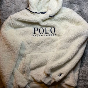 Vit fleece hoodie från Ralph Lauren - Mysig vit fleece hoodie från Ralph Lauren med broderad logga på bröstet och en liten broderad detalj på ärmen. Perfekt för kyliga dagar med sin fluffiga och varma design. Har en stor ficka framtill och justerbar huva.