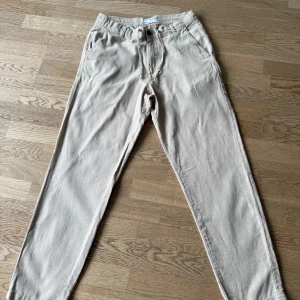 Beige byxor från Selected Homme - Snygga beige byxor från Selected Homme med elastisk midja och snörning. Byxorna har en avslappnad passform och är perfekta för en stilren look. De har både fram- och bakfickor med knappdetaljer.