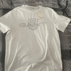 Vit t-shirt från GANT - Säljer en stilren vit t-shirt från GANT med deras ikoniska logga tryckt på bröstet. T-shirten har en normal passform och är kortärmad, perfekt för en avslappnad look. Är 190 och tycker den passar perfekt. Den är helt ny.