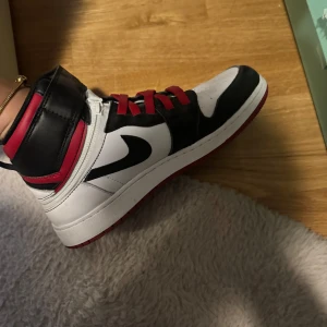 Nike Air Jordans i svart, vitt och rött - Snygga Nike Air Jordans i en klassisk färgkombination av svart, vitt och rött. Skorna har en hög design med snörning och den ikoniska Nike-loggan på sidan. Perfekta för att ge din outfit en sportig touch.