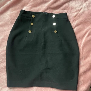 Svart kjol med guld knappar - Snygg svart kjol med dekorativa guldknappar framtill och en diskret slits bak. Kjolen har en dragkedja i ryggen för enkel på- och avtagning. Perfekt för en stilren look🖤🤍