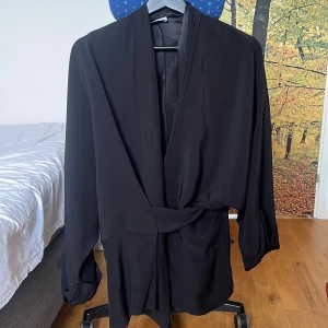 Svart kavaj från Rodebjer - Säljer en elegant svart kavaj från Rodebjer i 100% viskos. Kavajen har en stilren design med ett bälte i midjan och långa ärmar. Perfekt för en chic look.