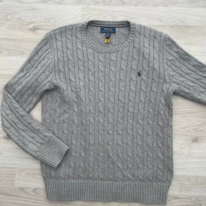 Ralph Lauren Kabelstickad  - Säljer en stilren grå stickad tröja från Ralph Lauren med kabelstickat mönster och broderad logotyp på bröstet. Tröjan har långa ärmar och rund halsringning. Perfekt för kyligare dagar! Den är i nytt skick och finns inga som helst defekter. Det är storlek 14-16 vilket motsvarar S.