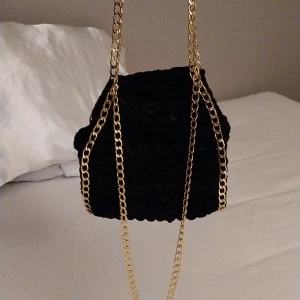 Svart virkad väska med guldkedja - Snygg svart virkad väska i med en elegant guldkedja. Perfekt för att ge en lyxig touch till din outfit. Väskan har en unik form och är en riktig statement piece.