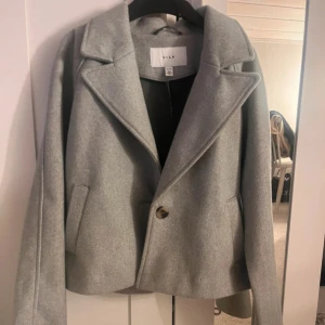 Grå kappa från Vila - Säljer en stilren grå kappa från Vila med en klassisk krage och en knapp framtill. Jackan har långa ärmar och en elegant design som passar perfekt för både höst och vår.