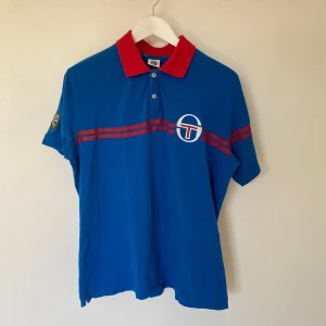 Blå pikétröja från Sergio Tacchini - Snygg blå pikétröja från Sergio Tacchini med röd krage och två knappar. Tröjan har ett tryck med logotyp på bröstet och texten 'Monte-Carlo' på ryggen. Storleken är XL men det passar som L.