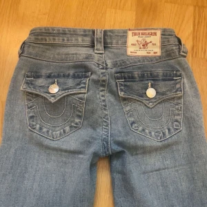 Blå bootcut jeans från True Religion - Snygga blå bootcut jeans från True Religion med klassiska femficksdesign och dekorativa sömmar på bakfickorna. Jeansen har en ljus tvätt och knappar med logotypdetaljer. Pris går alltid att diskuteras💓 Jag sydde upp byxorna i butik. I byxstorlek  har jag 152. Jag är oxå runt 150 och de passar perfekt på mej! Jag säljer för att de inte används och jag letar efter en annan färg 💕