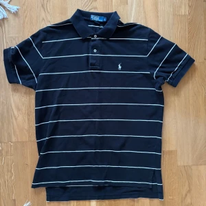 Svart och vit randig pikétröja från Ralph Lauren - Säljer en klassisk svart pikétröja från Ralph Lauren med vita ränder. Tröjan har korta ärmar och en broderad logotyp på bröstet. Perfekt för en stilren look.