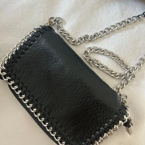 Svart axelväska från Tiamo - Snygg svart axelväska från Tiamo i skinn med en  kedjedetalj runt kanterna.  Säljer för 250kr använd sparsamt 