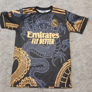 Svart och guld Real Madrids fotbollströja - Säljer en svart fotbollströja från Adidas med ett coolt drakmönster i guld. Tröjan har korta ärmar och Real Madrid-logga på bröstet. Perfekt för fotbollsfans som vill sticka ut på planen! 🐉⚽