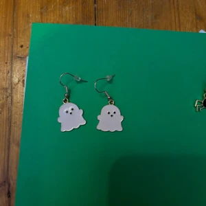 Spökeformade örhängen - Charmiga örhängen i form av små vita spöken med svarta ögon. Perfekta för att ge en lekfull touch till din outfit. De hänger från en enkel krok och är lätta att bära.
