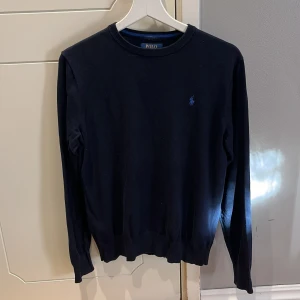 Mörkblå tröja från Polo Ralph Lauren - Säljer en stilren mörkblå tröja från Polo Ralph Lauren i 100% bomull. Tröjan har en klassisk design med rund hals och ett broderat logotypmärke på bröstet. Perfekt för en avslappnad och elegant look. Storlek: XL/TG (18-20) (Barn storlek)