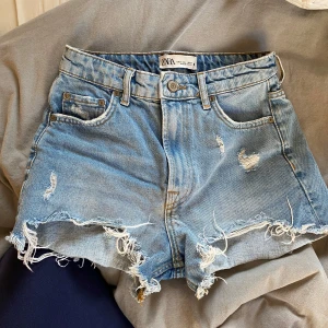 Blå jeansshorts från Zara - Säljer ett par coola blå jeansshorts från Zara med slitna detaljer och fransiga kanter. Perfekta för en avslappnad sommarlook. De har en klassisk femficksdesign och knappgylf. Passar perfekt för varma dagar! 🌞