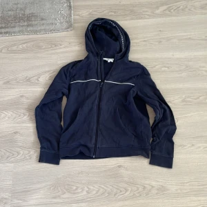 Mörkblå huvtröja från Hugo Boss - Säljer en snygg mörkblå huvtröja från Hugo Boss med dragkedja och huva. Tröjan har en diskret logga på bröstet och en dekorativ rand över bröstet. Perfekt för en casual look.