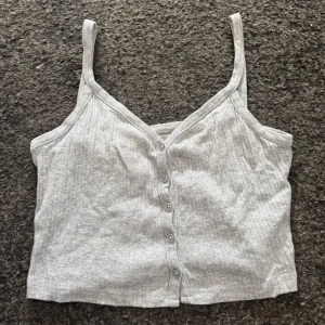 Grå ribbad linnetopp från Brandy Melville - Säljer en grå ribbad linnetopp från Brandy Melville med knappar framtill. Toppen har smala axelband och en v-ringad design. Perfekt för en avslappnad stil. Så skönt material!!