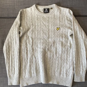 Grå stickad tröja från Lyle & Scott - Säljer en stilren grå stickad tröja från Lyle & Scott med ett broderat gult emblem på bröstet. Tröjan har ett klassiskt kabelstickat mönster och långa ärmar. 