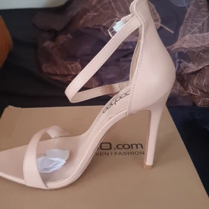 Beige klackskor från Boohoo - Snygga beige klackskor från Boohoo med öppen tå och smal rem över foten. Skorna har en elegant ankelrem med spänne och hög klack för en stilren look.