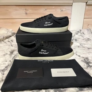Svarta sneakers från Saint Laurent - Snygga svarta sneakers från Saint Laurent med vit text på sidan. Skorna har en klassisk design med snörning och en vit sula. Perfekta för en stilren look.