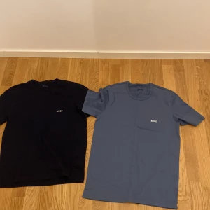 Svart och blå t-shirt från Boss - Säljer två stilrena t-shirts från Boss i storlek M. En svart och en blå, båda med korta ärmar och klassisk rund hals. Perfekta för en avslappnad look. Boss-loggan är diskret placerad på bröstet.