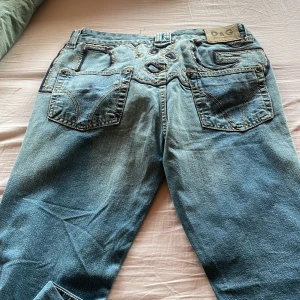 Blå jeans från D&G - Snygga blå jeans från D&G med unika broderade detaljer på bakfickorna. Jeansen har en klassisk femficksdesign och knappar i midjan. Perfekta för en stilren look.