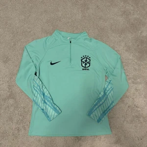 Ljusblå långärmad träningströja från Nike BRAZIL - Snygg ljusblå träningströja från Nike med Brasiliens landslagsmärke på bröstet. Tröjan har en halv dragkedja framtill och ett coolt mönster på ärmarna. Perfekt för fotbollsträning eller andra sportaktiviteter. Använd endast tre gånger och är i bra skick och vill sälja den. Storlek M