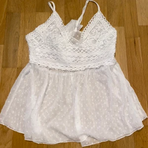Vit spetslinne med broderad detalj - Säljer en söt vit spetslinne med broderade detaljer och en v-ringad design. Toppen har tunna axelband och en knytning i ryggen. Den är perfekt för sommarens varma dagar och har en luftig, lätt känsla.