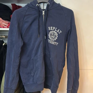 Blå hoodie från Replay - Säljer en blå hoodie från Replay med dragkedja och tryck på både framsidan och baksidan. Tröjan har en justerbar huva med snören och praktiska fickor framtill. Perfekt för en avslappnad stil.