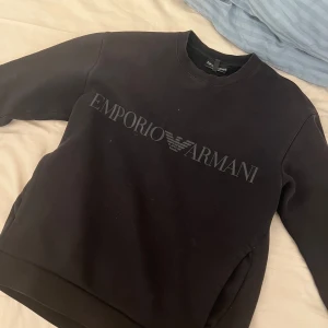 Svart sweatshirt från Emporio Armani - Säljer en stilren svart sweatshirt från Emporio Armani med deras ikoniska logga tryckt över bröstet. Tröjan har en klassisk rund hals och långa ärmar. Perfekt för en avslappnad och trendig look.