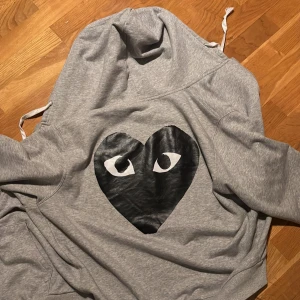 Grå hoodie från Comme des Garçons - Säljer en grå hoodie från Comme des Garçons med det ikoniska svarta hjärtat med ögon på bröstet och ett mindre på ärmen. Perfekt för en stilren och avslappnad look.