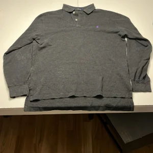 Grå pikétröja från Polo Ralph Lauren - Säljer en klassisk grå pikétröja från Polo Ralph Lauren med lång ärm och knappar i halsen. Tröjan har en broderad lila logotyp på bröstet. Perfekt för en stilren look.