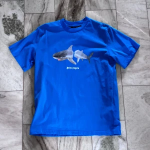 Blå Palm Angels Shark T-Shirt - Säljer en blå Palm Angels Shark t-shirt med ett hajmotiv på framsidan och Palm Angels-logga på baksidan. Storlek M.