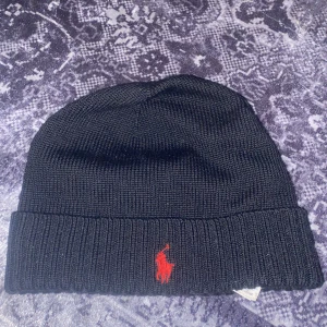 Svart mössa - Snygg svart stickad mössa med röd broderad logga framtill. Perfekt för att hålla värmen under kyliga dagar. Klassisk design som passar till det mesta.