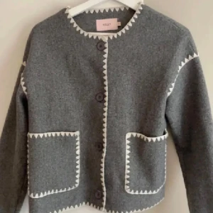 Grå kavaj från Nelly  - Säljer en stilren grå kavaj från Nelly med vita detaljer längs kanterna. Kavajen har knappar framtill och två praktiska fickor. Perfekt för en elegant look. Storlek xs, passar ej mig så fler bilder finns ej