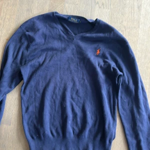 Mörkblå tröja från Polo Ralph Lauren - Säljer en stilren mörkblå tröja från Polo Ralph Lauren med v-ringning och det klassiska logotypbroderiet i orange på bröstet. Perfekt för en avslappnad men ändå elegant look.
