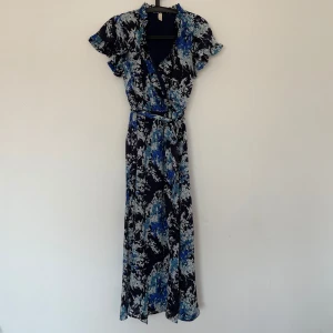 Blommig långklänning från bubbleroom  - Snygg klänning med blommigt mönster i blått och vitt på en mörk bas. Klänningen har ärmar med volangdetaljer. Perfekt för en somrig look!