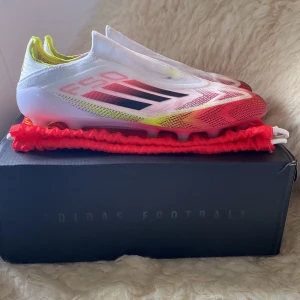 Adidas F50 Elite LL AG Fotbollsskor - Snygga Adidas F50 Elite LL AG fotbollsskor i vitt med inslag av svart och gul. Skorna har en strömlinjeformad design med en färggradient från vit till rosa på sulan. Perfekta för konstgräsplaner och ger ett bra grepp och kontroll. Använt fåtal gånger. Storlek 45 1/3. Det kommer nya sulor och fotbollspåse som man kan lägga skorna i. Kvitto finns också! Köpt på unisport i Stockholm.