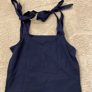 Linne med rosetter - Säljer en stilren mörkblå linnetopp från Holly & White. Toppen har knytbara axelband som ger en söt detalj och är perfekt för varma sommardagar. Använd en gång och är i nyskick.