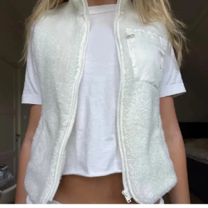 Vit fleeceväst med dragkedja - Säljer en mysig vit fleeceväst med dragkedja framtill. Västen har en praktisk bröstficka och är perfekt för lager-på-lager under kyligare dagar.