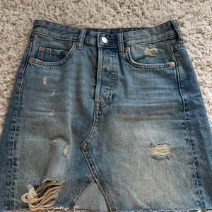 Jeanskjol med slitningar - Säljer en cool jeanskjol med slitningar och fransig nederkant. Kjolen har en klassisk femficksdesign och en knappgylf. Perfekt för en avslappnad stil.