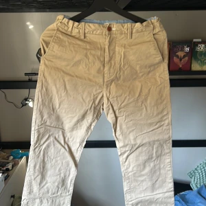 Beige chinos från Gant - Snygga beige chinos från Gant med normal passform. Byxorna har knappar och bälteshällor, vilket ger en klassisk look. Perfekta för en stilren vardagsoutfit.