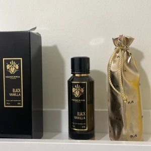 Black Vanilla Eau de Parfum från Mancera - Elegant och lyxig parfymflaska med doften Black Vanilla från Mancera. Flaskan är svart med gulddetaljer och kommer i en stilren svart kartong samt en glansig guldfärgad påse med dragsko. Perfekt för den som älskar en djup och sensuell doftupplevelse.