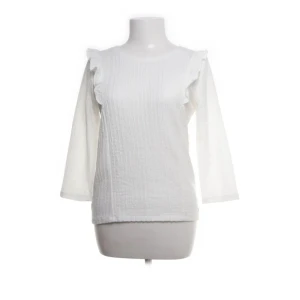 Vit blus  - Säljer en stilren vit blus med volangdetaljer på axlarna. Blusen har trekvartsärmar och en ribbad struktur som ger en elegant touch. Perfekt för att lyfta din vardagslook eller för speciella tillfällen.