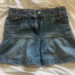 Jeanskjol med volang - Snygg jeanskjol i blå denim med volangdetaljer. Kjolen har en klassisk femficksdesign och stängs med dragkedja och knapp framtill. Perfekt för en avslappnad stil.