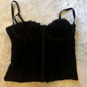 Svart korsett med spetsdetaljer - Snygg svart korsett med spetsdetaljer och axelband. Den har en elegant snörning bak och är perfekt för en stilfull look. Passar bra till både fest och vardag. Använd fåtal gånger. Stolek Xs/S