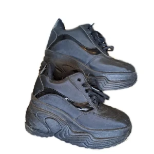 Svarta sneakers med platåsula - Snygga svarta sneakers med hög platåsula och snörning. Skorna har en kombination av glansiga och matta detaljer som ger en cool look. Perfekta för att ge lite extra höjd och stil till din outfit.