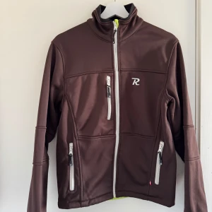 Brun jacka från Rumble Clothing - Säljer en brun jacka från Rumble Clothing med dragkedja och flera praktiska fickor. Jackan har en broderad logga på bröstet och är perfekt för kyligare dagar. Den har långa ärmar och en bekväm passform. (Katt o kanin finns i hemmet)