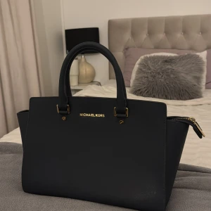 handväska från Michael Kors - Elegant mörk handväska från Michael Kors i skinn med guldfärgade detaljer och dragkedja. Perfekt för att bära dina dagliga nödvändigheter med stil. Väskan har ett rymligt inre med flera fack för enkel organisering.